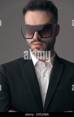 Closeup of confident fashion model wearing suit and sunglasses with hands crossed while standing on gray studio background Stock Photo