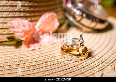 Composition of two wedding rings or bands and engagement ring with diamond on blurred background of straw hat, pink flower and silver bracelet Stock Photo