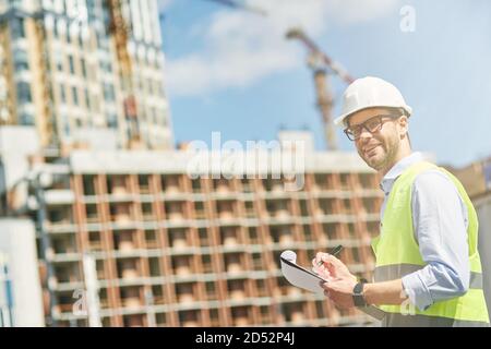Young civil engineer or construction supervisor wearing helmet smiling at camera while inspecting building site and making some notes Stock Photo