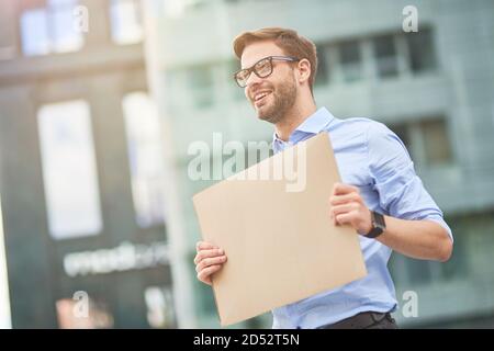 Portrait of a young cheerful man, male activist wearing blue shirt and eyeglasses holding empty sign board and smiling while protesting outdoors Stock Photo