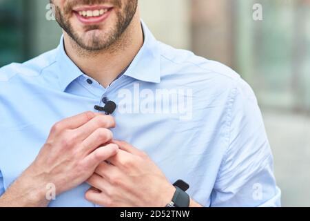 Young handsome man, male blogger adjusting small lavalier microphone on his shirt and smiling while standing outdoors Stock Photo