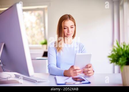 Portrait of happy young businesswoman holding digital tablet in her hands and working while sitting at office desk in front of laptop. Stock Photo