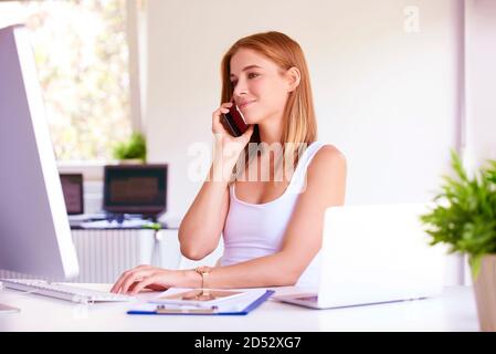 Beautiful young businesswoman talking with someone on her mobile phone while sitting in front of computer. Stock Photo