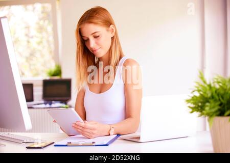 Smiling young woman working at the office. Beautiful businesswoman sitting at office desk and using digital tablet. Stock Photo