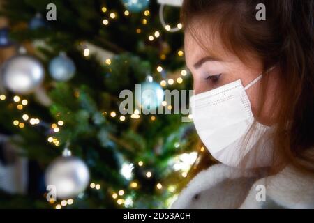 Portrait of a young woman wearing protective face mask and looking sad for Covid-19 with Christmas tree on the background, coronavirus and Christmas Stock Photo