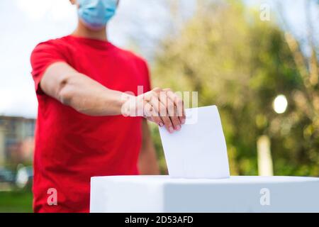 Man with face mask putting his vote into ballot box on table outside. Stock Photo