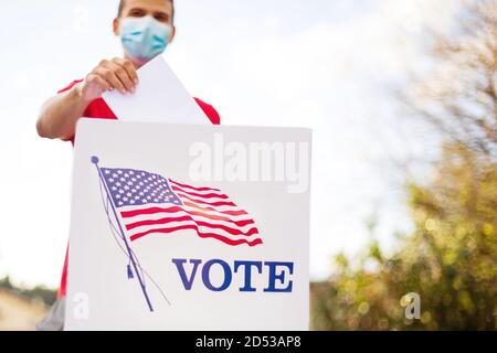 Man with face mask putting his vote into ballot box on table outside. Stock Photo