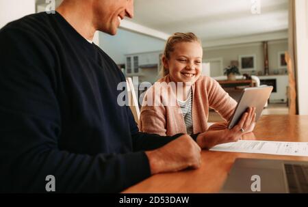 Father and daughter sitting at table using a digital tablet together at home. Girl looking at tablet computer in hand of her father at home. Stock Photo
