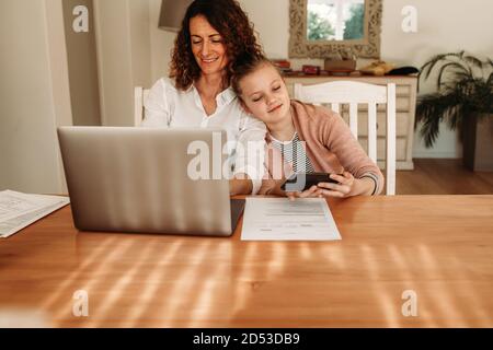 Girl using mobile phone while sitting close to her mother working on a laptop. Woman working from home sitting with her daughter using a smart phone. Stock Photo