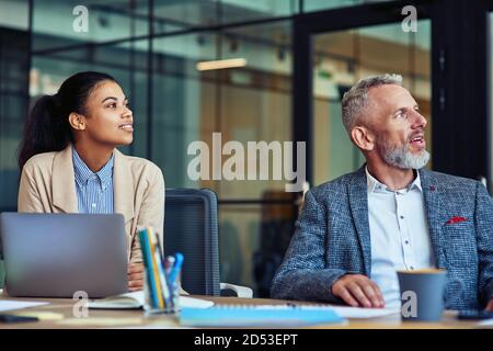 Business people at work. Young mixed race woman and her mature colleague looking away while sitting at desk and working together in the mode office Stock Photo