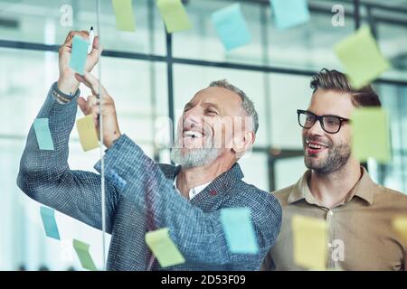 Two happy male coworkers putting colorful sticky notes on a glass wall and smiling while standing in the modern office Stock Photo