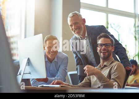 Happy business people. Three cheerful men looking at camera and smiling while sitting at desk and using PC, working on project together in the modern Stock Photo