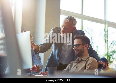 Business people sitting at desk in the modern office, looking at PC screen and discussing project, confident mature man explaining something to his Stock Photo