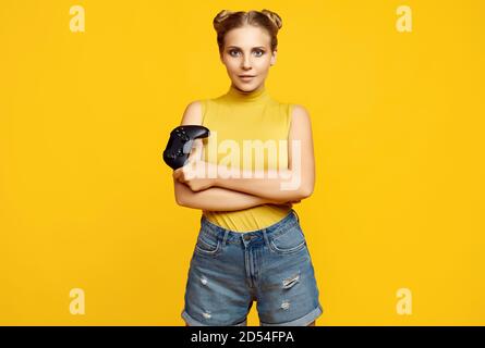 Portrait of gorgeous happy blonde gamer girl playing video games using joystick on yellow background in studio Stock Photo