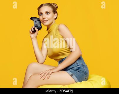 Portrait of gorgeous happy blonde gamer girl playing video games using joystick on yellow background in studio Stock Photo