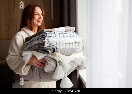 Young woman with beautiful smile in warm beige robe is leaning on stack of folded bed sheets Stock Photo