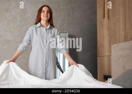 Happy woman with long red hair is folding clean bedding set on the bed of room with copy space. Stock Photo