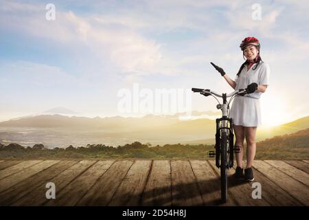 An Asian woman with a bicycle helmet standing beside her bicycle on a wooden path with a landscape background Stock Photo