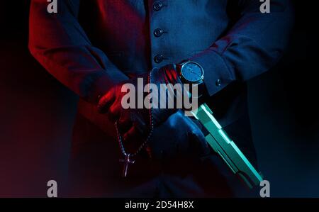 Photo of a male mafia criminal killer in suit and leather gloves holding a gun with cross on black background. Stock Photo