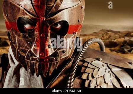 Photo of a post apocalyptic raider warrior in leather jacket with metal armor and steel mask with red cross painting standing in desert wasteland. Stock Photo