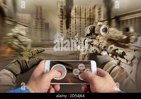 First person view photo of a male hand in jeans shirt holding smartphone and playing war battlefield shooter game. Stock Photo