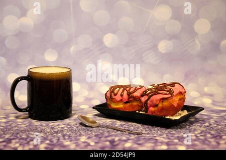 Glass cappuccino mug with foam and rectangular black plate with two donuts in pink glaze with chocolate and powder on a gray background. Close-up. Stock Photo