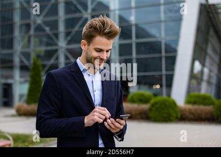 Handsome office worker holding smartphone and smiling. Happy young man using mobile phone apps, texting message, browsing internet, looking at Stock Photo