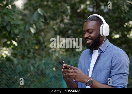 Portrait of smiling guy in earphones and mobile phone outdoors. Young Afro-American man in headphones listening music on smart phone using music app Stock Photo