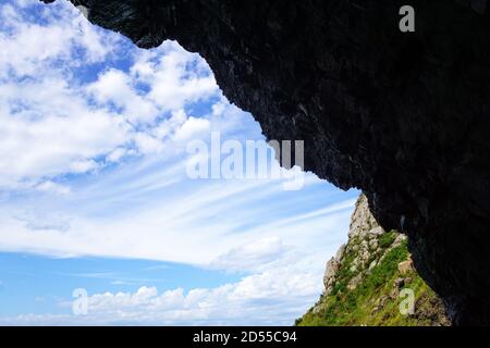 A beautiful view opens up through the hole from the cave: mountains, clear blue sky and white clouds. Dark wall above the exit. Inside is darkness, bu Stock Photo