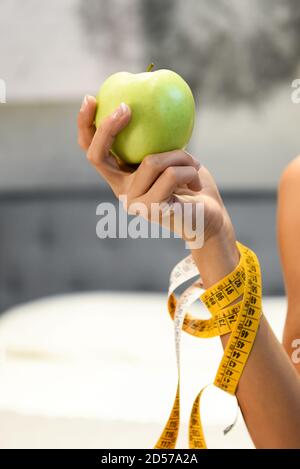 Woman holding a fresh healthy green apple in her hand with a tape measure wound round her arm in a diet and weight loss concept in close up Stock Photo