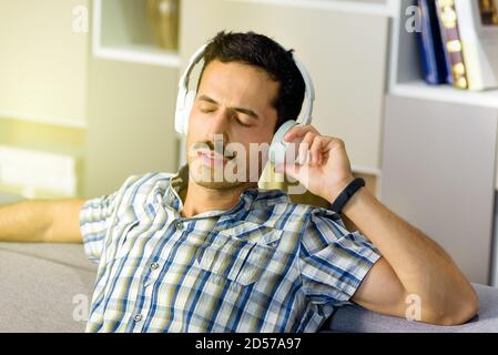 Blissful young man listening to music on stereo headphones as he relaxes on a sofa at home with eyes close immersed in the soundtrack Stock Photo