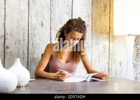 Attractive young woman working on crossword puzzles at home in a magazine seated at a table with copyspace Stock Photo