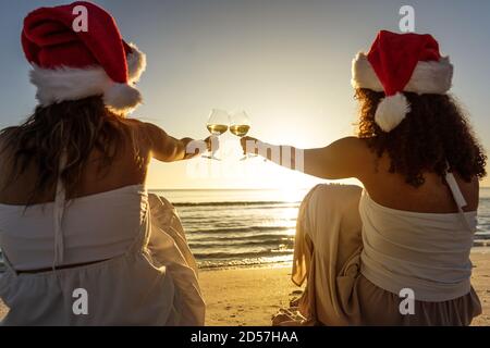 Rear view of two multiethnic best female friends wearing Santa hat sitting on the seashore and toasting with a glass of white wine in the sunset or da Stock Photo