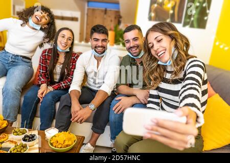 Group of mixed race friends making selfie celebrating at home with medical protection mask - Happy people indoor aperitif party at the Coronavirus new Stock Photo