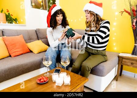 Female multi ethnic couple with Santa hat, celebrating at home giving gift with glasses of white wine - Caucasian blonde cute woman enjoying Christmas Stock Photo
