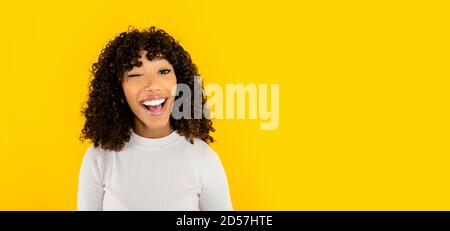 Black Hispanic curly beautiful young woman casual dressed winks looking at the camera smiling with open mouth on big yellow background copy space - On Stock Photo