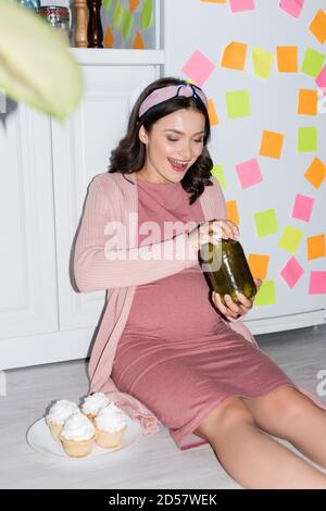 selective focus of joyful woman holding jar with canned cucumbers while sitting on floor near cupcakes Stock Photo