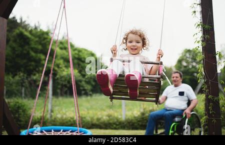 Small girl with senior grandfather in wheelchair playing in backyard garden, swinging. Stock Photo