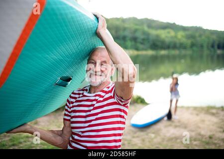 Senior man carrying paddleboard by lake in summer, looking at camera. Stock Photo
