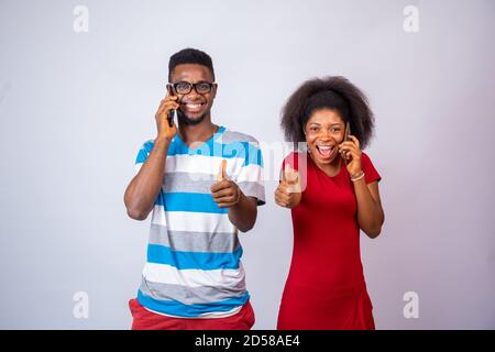 two excited young african people making phone calls, standing against a white background, give thumbs up Stock Photo