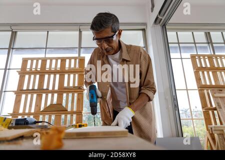 Asian senior man use electric drill on wooden board in carpentry workshop. Concept senior people doing hobby after retirement. Stock Photo