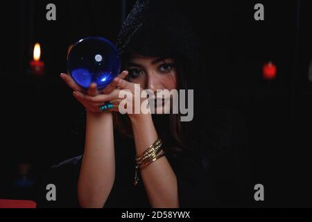 Portrait of Asian fortune teller in black hood holding blue crystal ball. Stock Photo