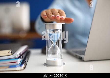 Woman holds her hand over hourglass at workplace in office closeup Stock Photo