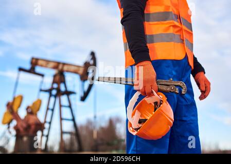 Cropped close-up snapshot of a man wearing blue overalls orange vest and gloves, holding a pipe wrench and a helmet on foreground, oil pump jack on background. Concept of petroleum industry. Stock Photo