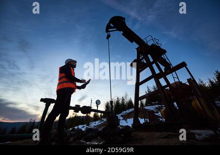 Low angle side view snapshot of petroleum engineer wearing orange vest and a helmet, looking at the oil rig and making some notes against beautiful evening sky, sunset Stock Photo