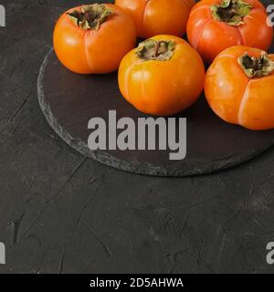 Group of fresh orange ripe persimmon fruits laying on round black slate on a dark background texture and copy space.  Stock Photo