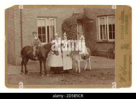 Original typical charming Victorian 1890's cabinet card of young children on a donkey and a pony outside a country house with the parlour maids, Victorian maids, Victorian servants, Worksop,  Nottinghamshire, England, U.K. circa 1894 Stock Photo