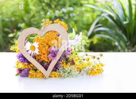 Bouquet of tansy, white daisy and thorny burdock wild summer flowers with wooden decorative heart Stock Photo
