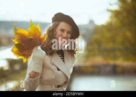 Hello november. happy trendy middle aged woman in beige trench coat with autumn yellow leaves outside in the city in autumn. Stock Photo