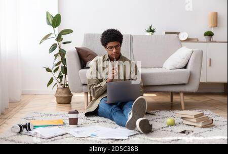 Focused black teen guy sitting on floor with study materials and laptop, at home Stock Photo